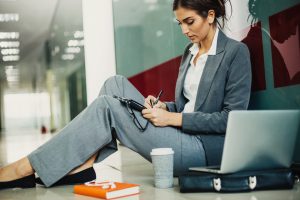 Young woman in business wear sitting on the floor next to a laptop and coffee cup, writing in a notebook, with copy space.